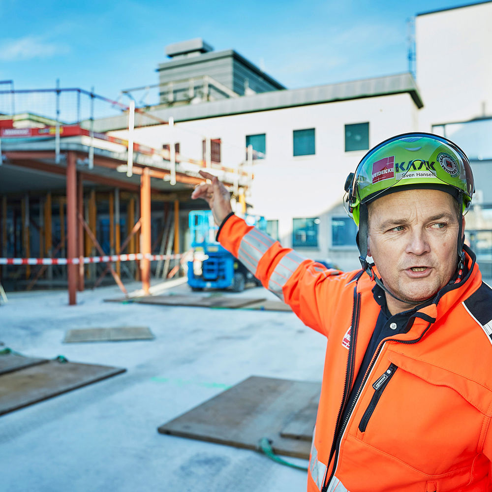 Mann med hjelm peker på tak inne på byggeplass. Foto.