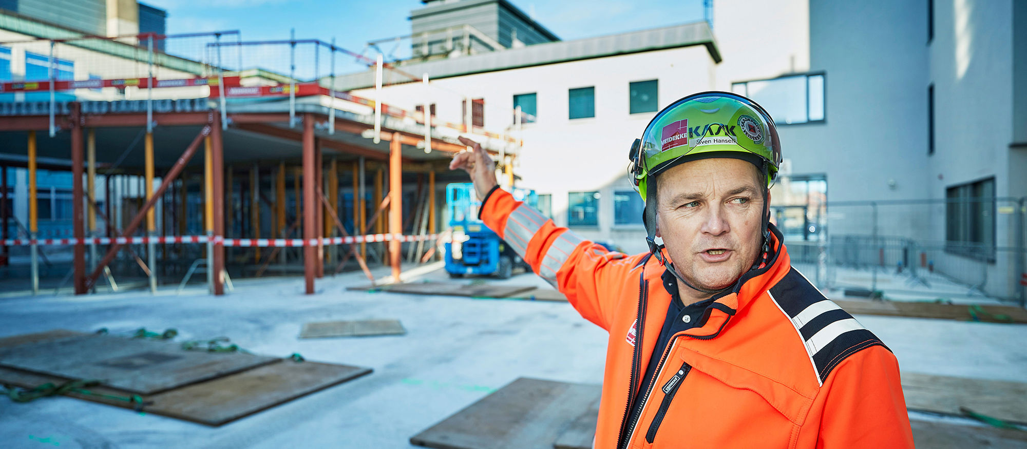 Mann med hjelm peker på tak inne på byggeplass. Foto.