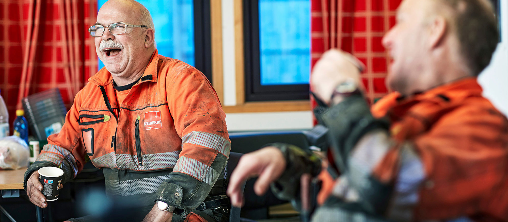 Two men lauging wearing high-vis clothing. Drinking coffee. Photo.